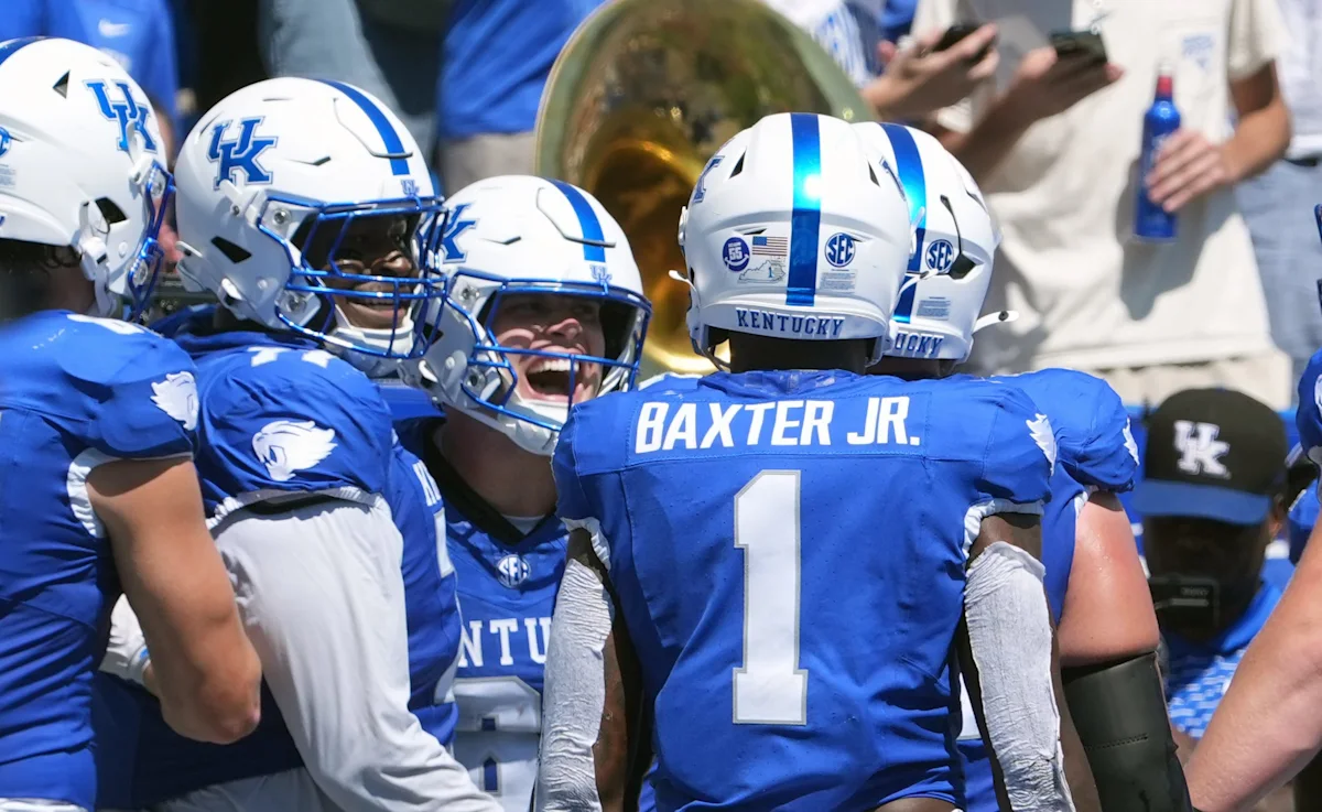 Kentucky football players in blue uniforms gather and celebrate on the field.