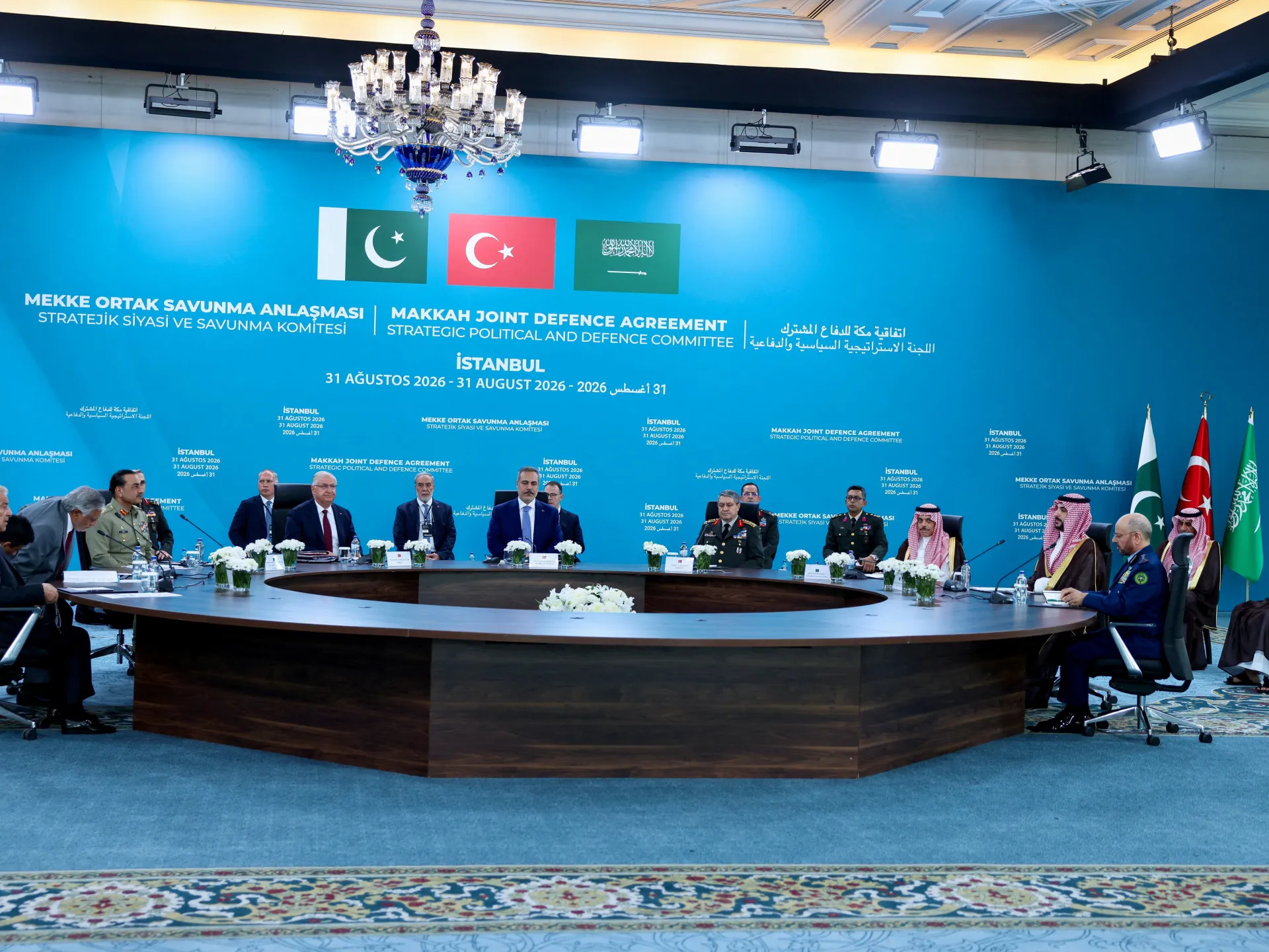 Officials and military representatives meet beneath Pakistani, Turkish and Saudi flags at a conference table.