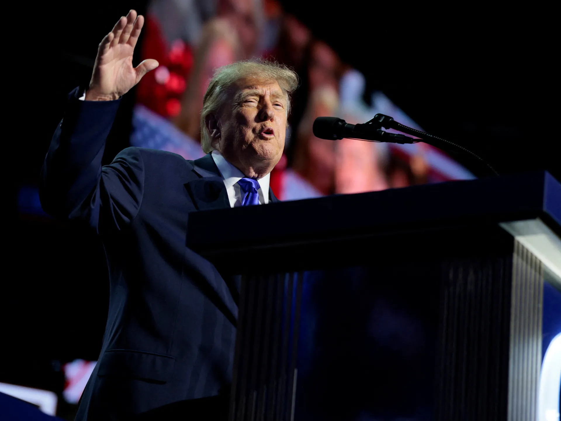 Donald Trump raises his hand while speaking at a podium with a microphone.