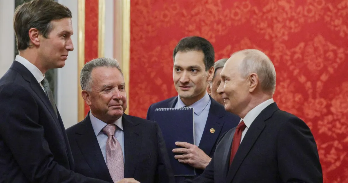 Four suited men converse indoors, with one holding a folder near a red patterned wall.