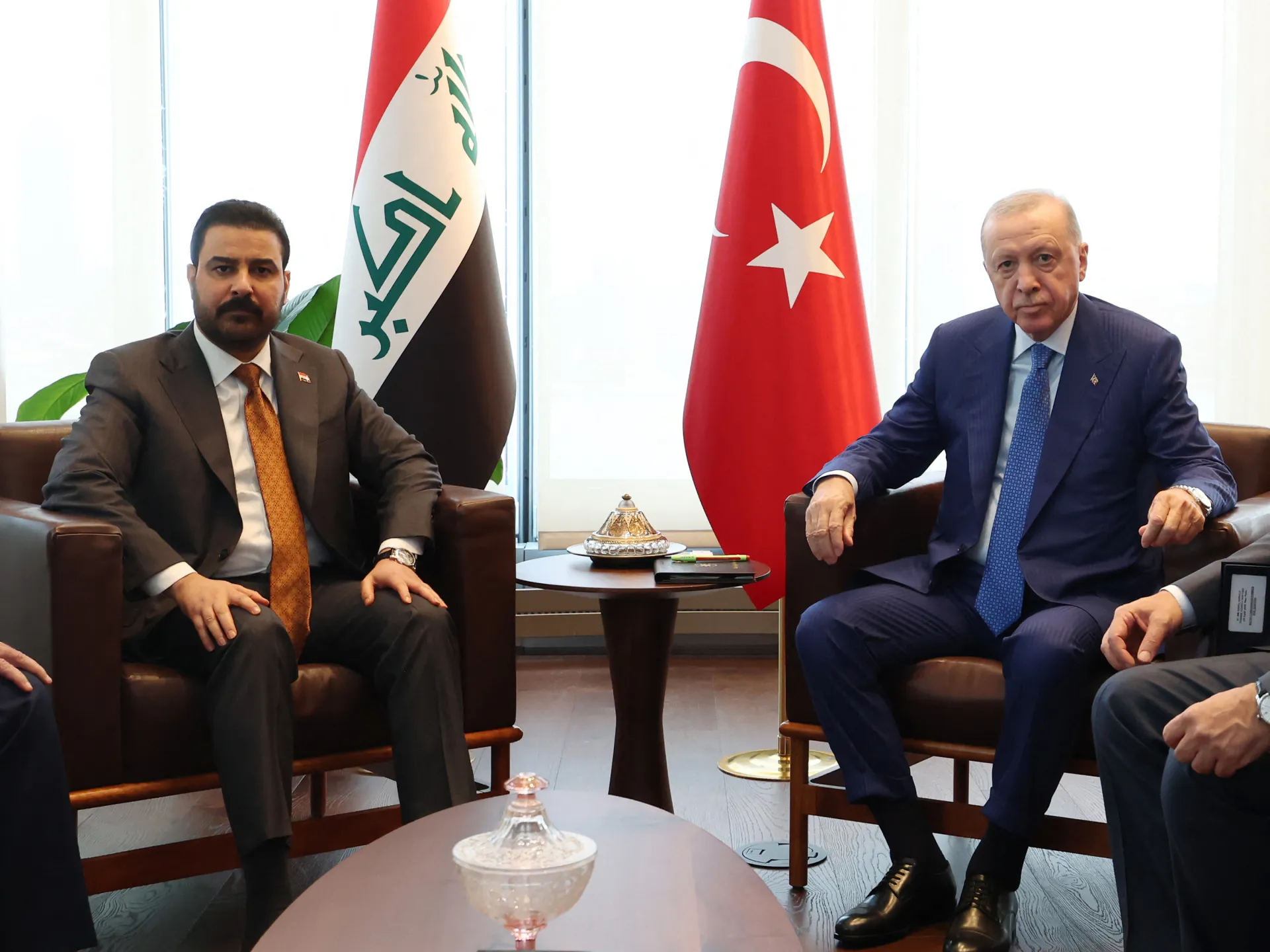 Two suited officials sit facing the camera between Iraqi and Turkish flags in a bright meeting room.