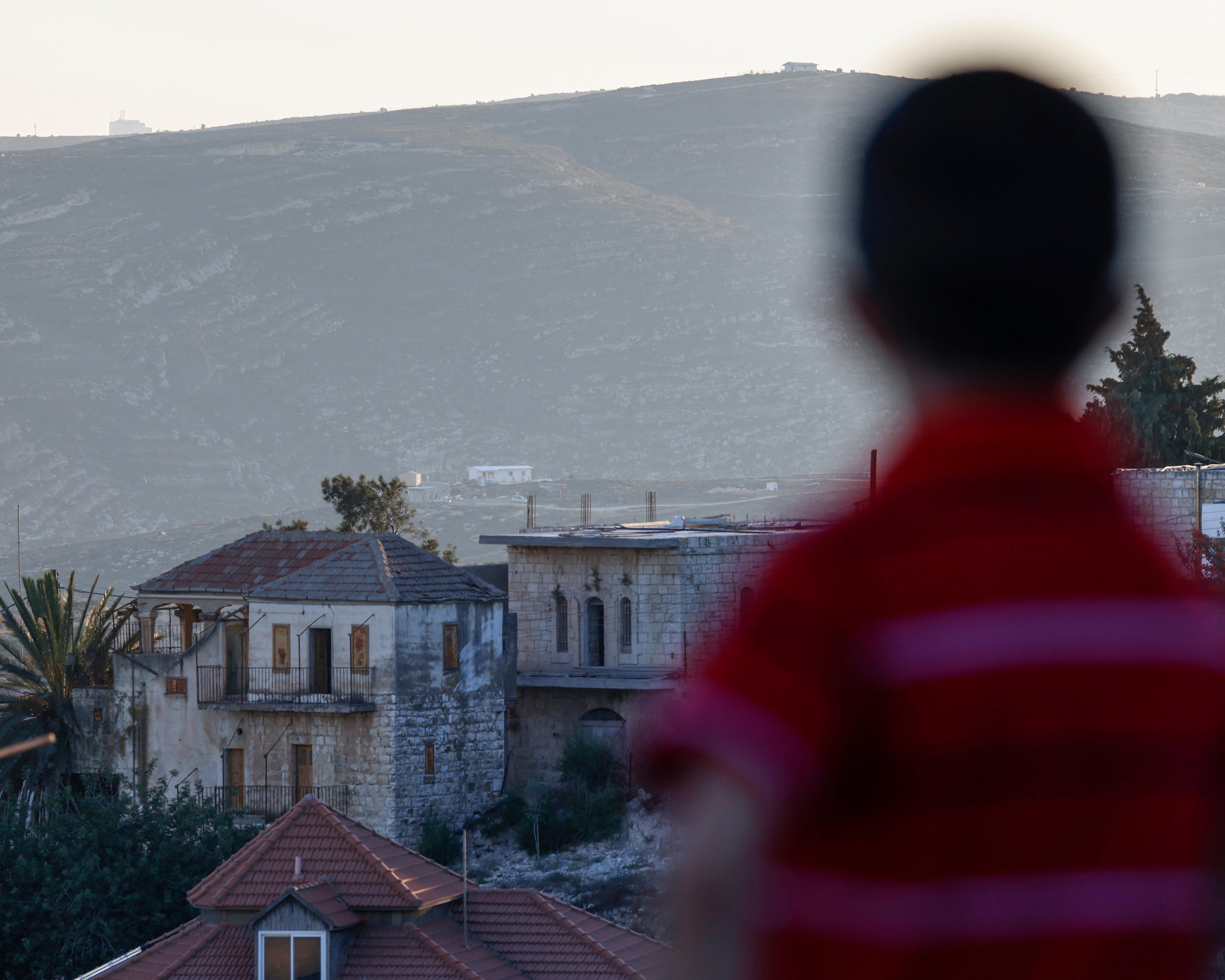 Blurred figure in red stands before stone homes and a barren hillside settlement.