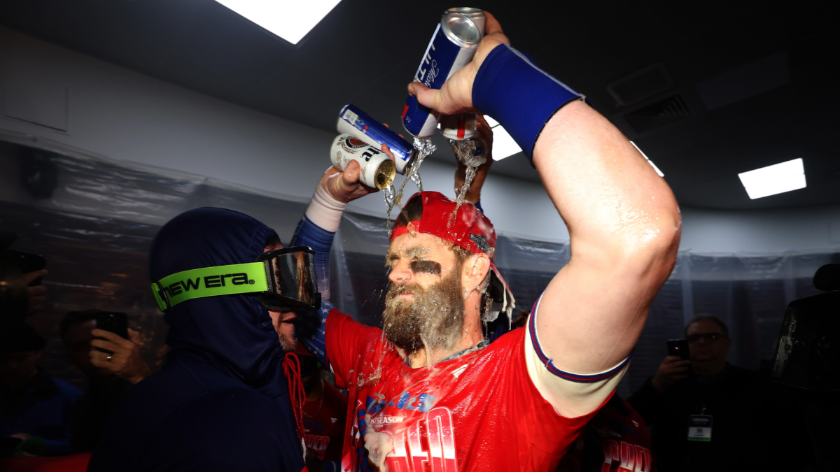 Bearded baseball player in a red jersey raises his arms as people pour drinks over him.