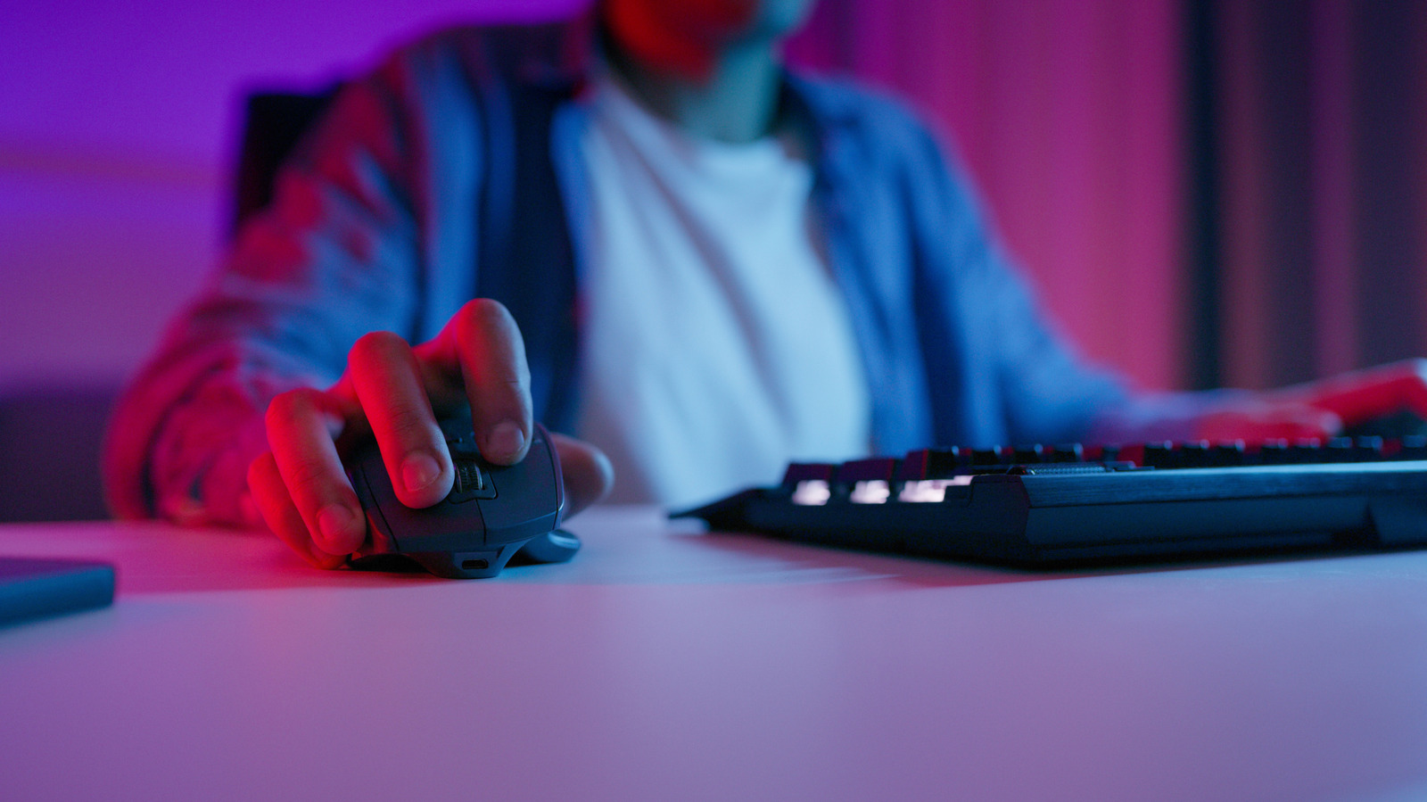 Close-up of a hand using a gaming mouse beside a keyboard under blue and pink lighting