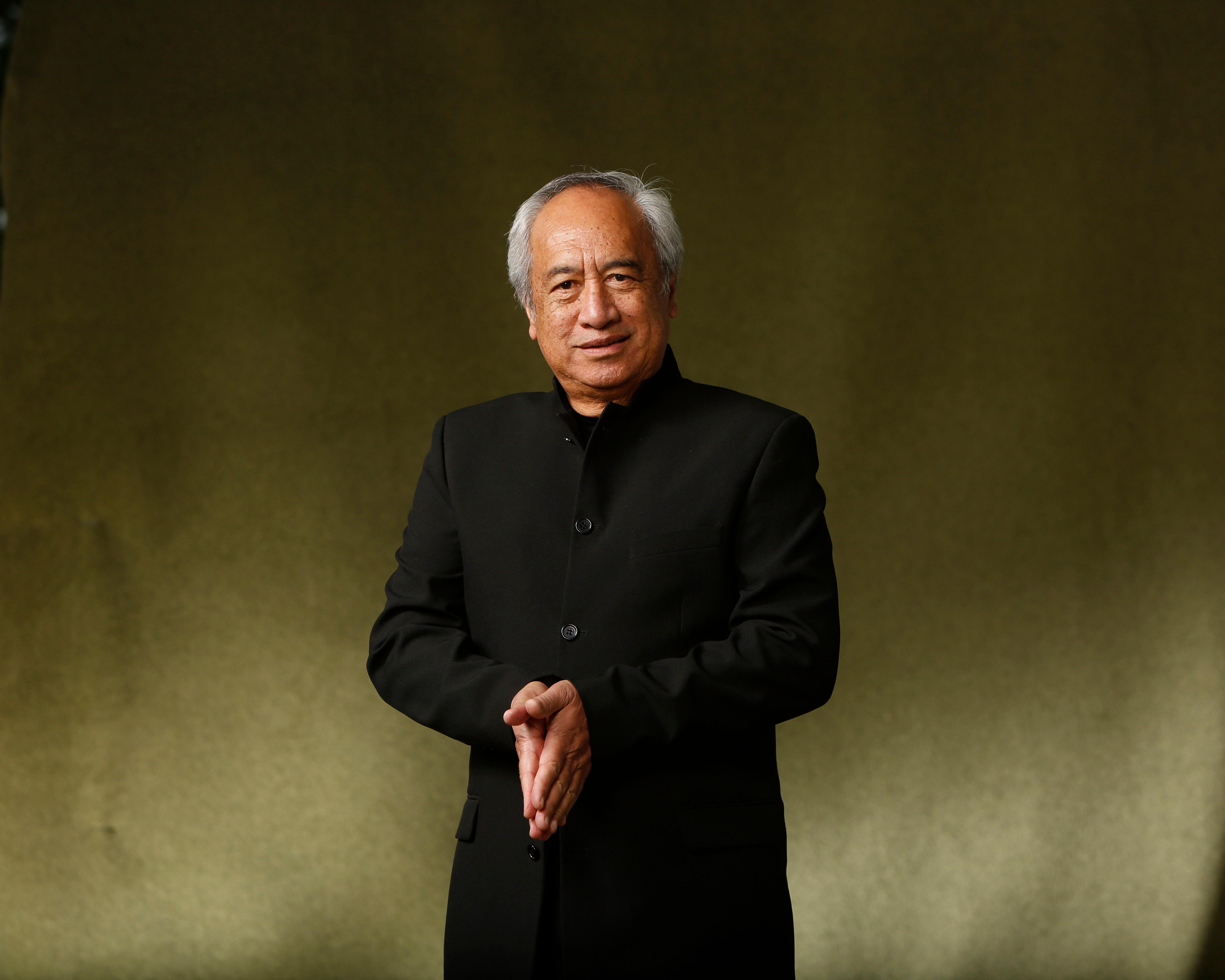 Older man with gray hair wearing a black jacket, standing against a mottled olive-brown backdrop.