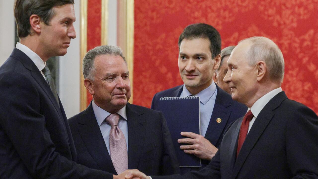 Four suited men converse indoors, with one holding a folder and another extending his hand.