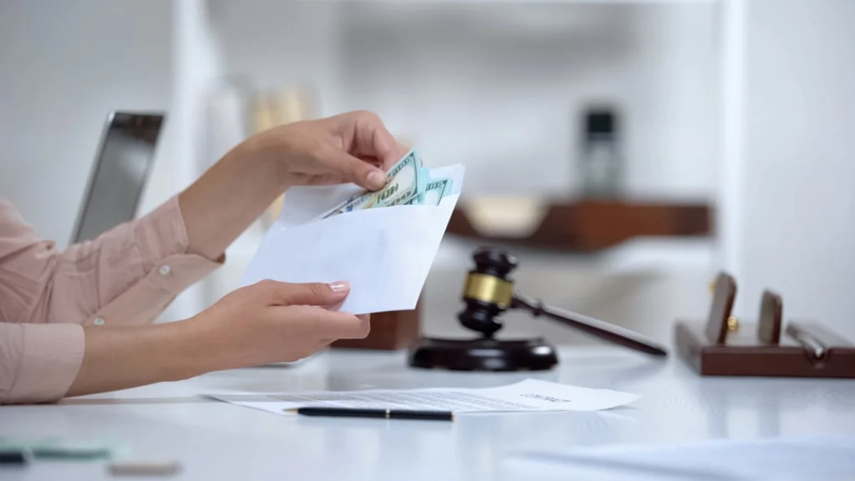 Person holding an envelope containing cash beside legal documents and a judge's gavel