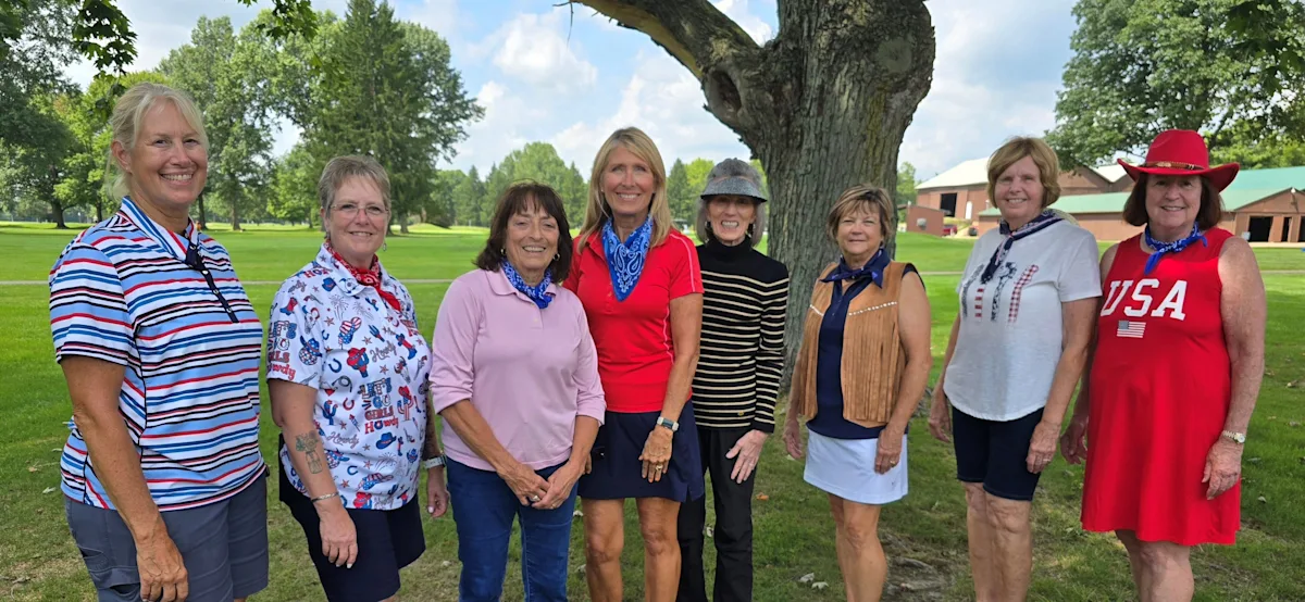 Nine women golfers pose together on a grassy lawn beneath a large tree.