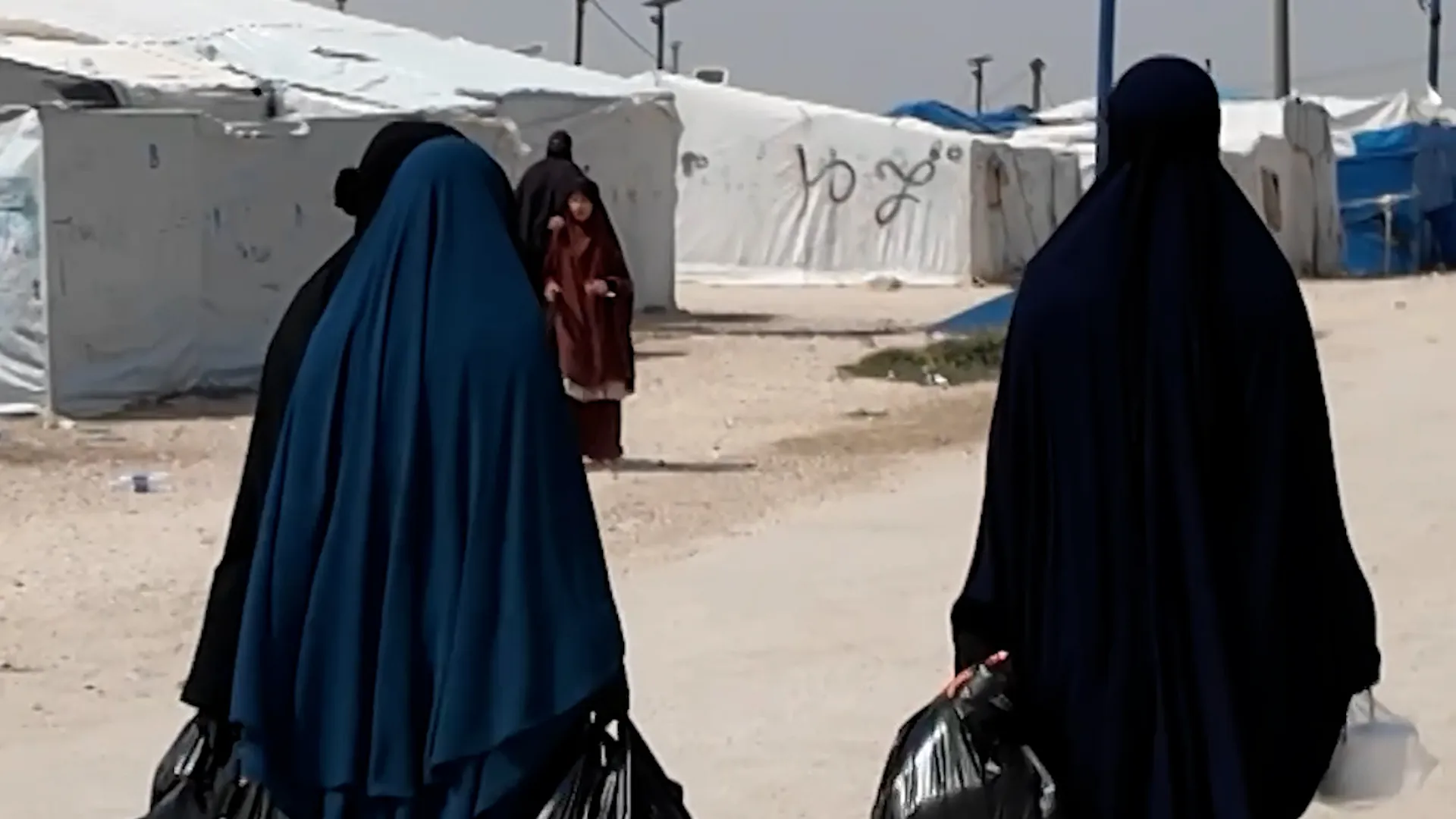 Two women in full-body coverings walk through a dusty camp past tents and a child.