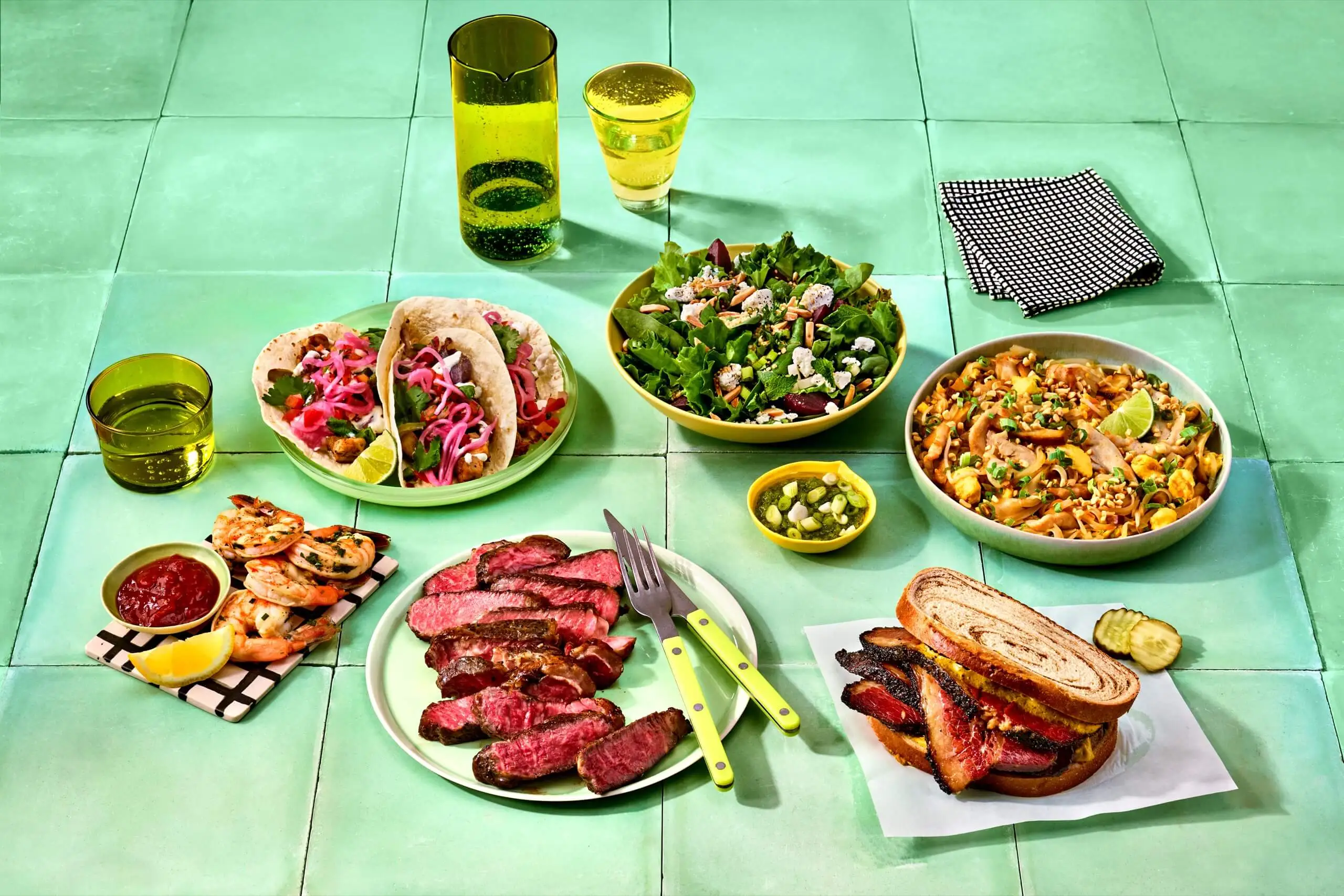 Assorted tacos, salad, noodles, grilled meats, shrimp, and sandwiches arranged on a turquoise tiled table.