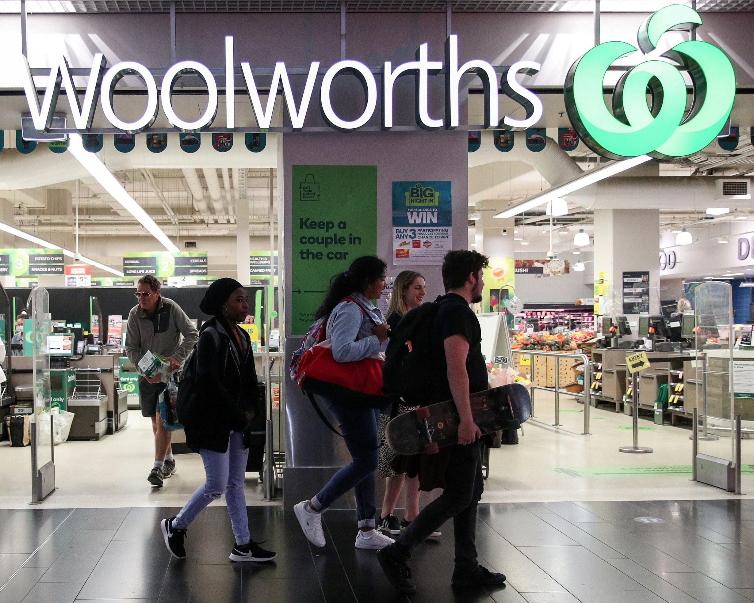 Shoppers walk past a Woolworths supermarket entrance and checkout area carrying bags and a skateboard.