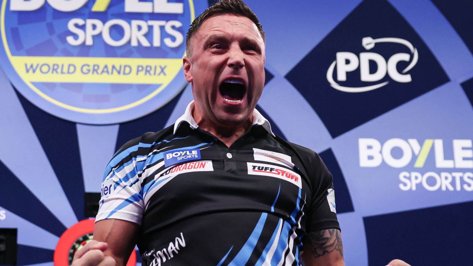 Darts player in a black and blue shirt shouts in front of PDC and World Grand Prix branding.