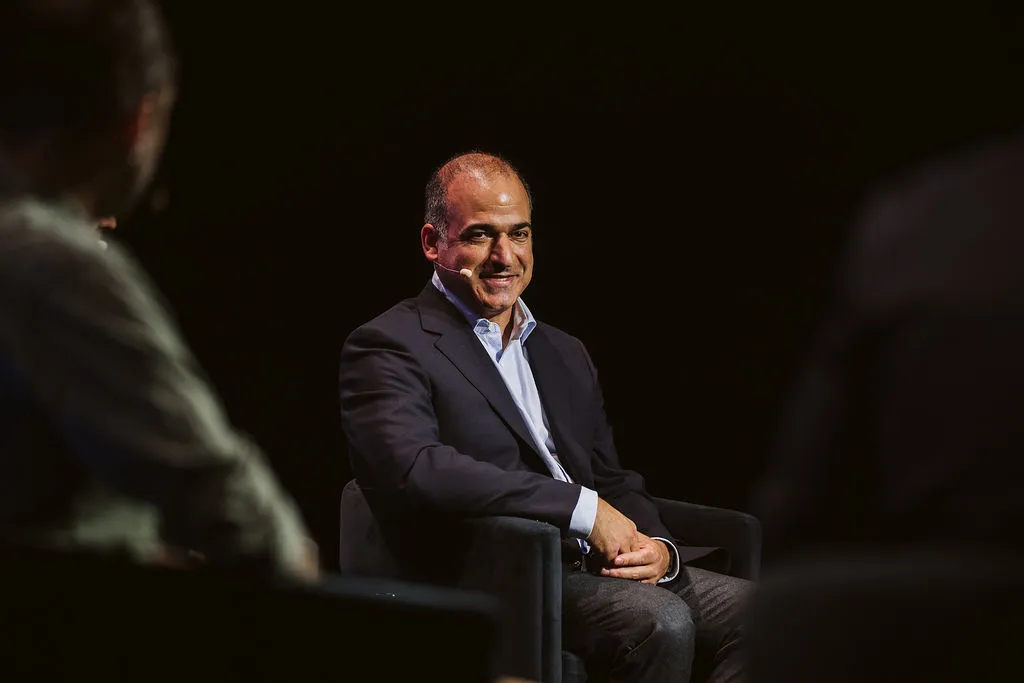 Man in a suit sits onstage beneath a spotlight, framed by two shadowed audience figures.