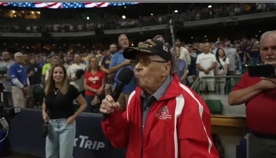 Elderly man in red jacket and cap sings into a microphone before a cheering crowd.