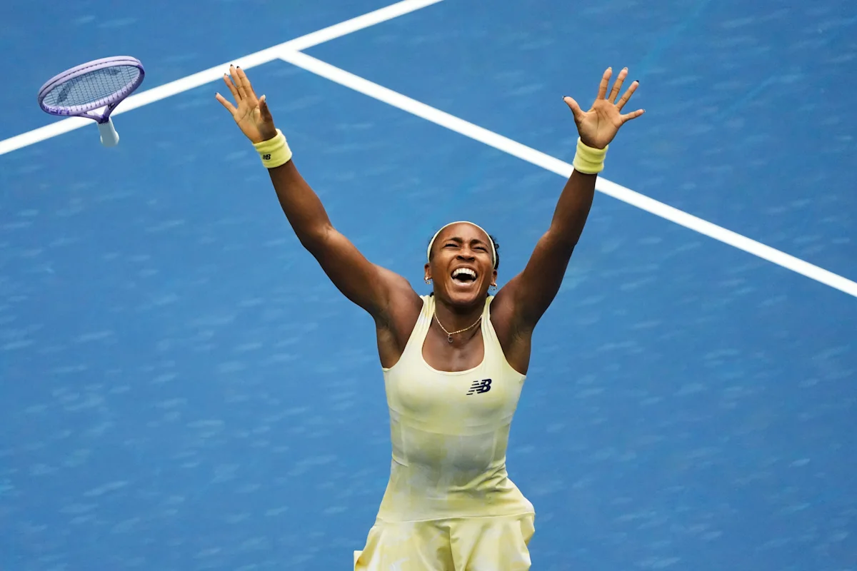 Tennis player celebrates with arms raised on a blue court, racket airborne nearby.