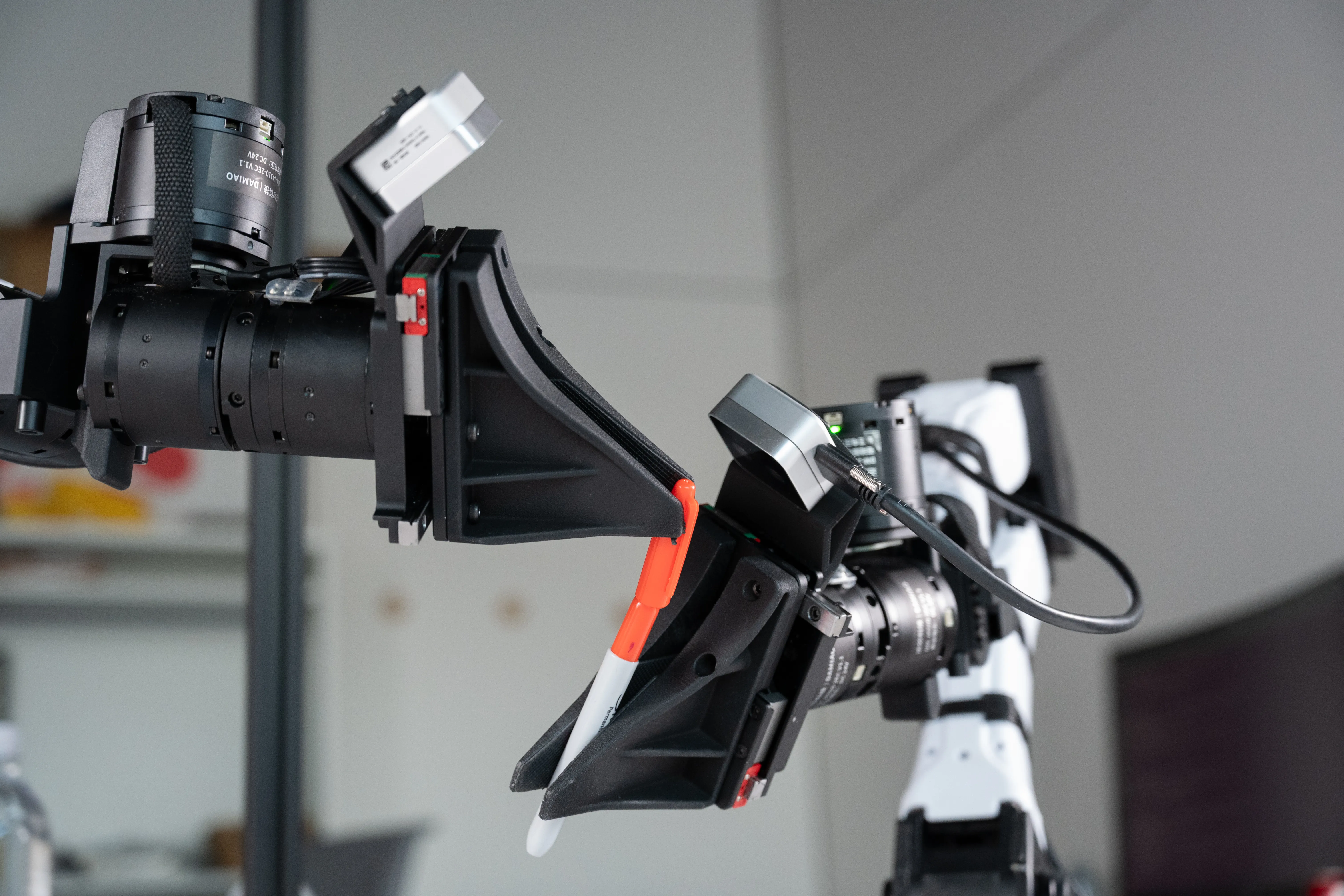 Two camera-equipped robotic grippers hold a marker in a laboratory or workshop setting.