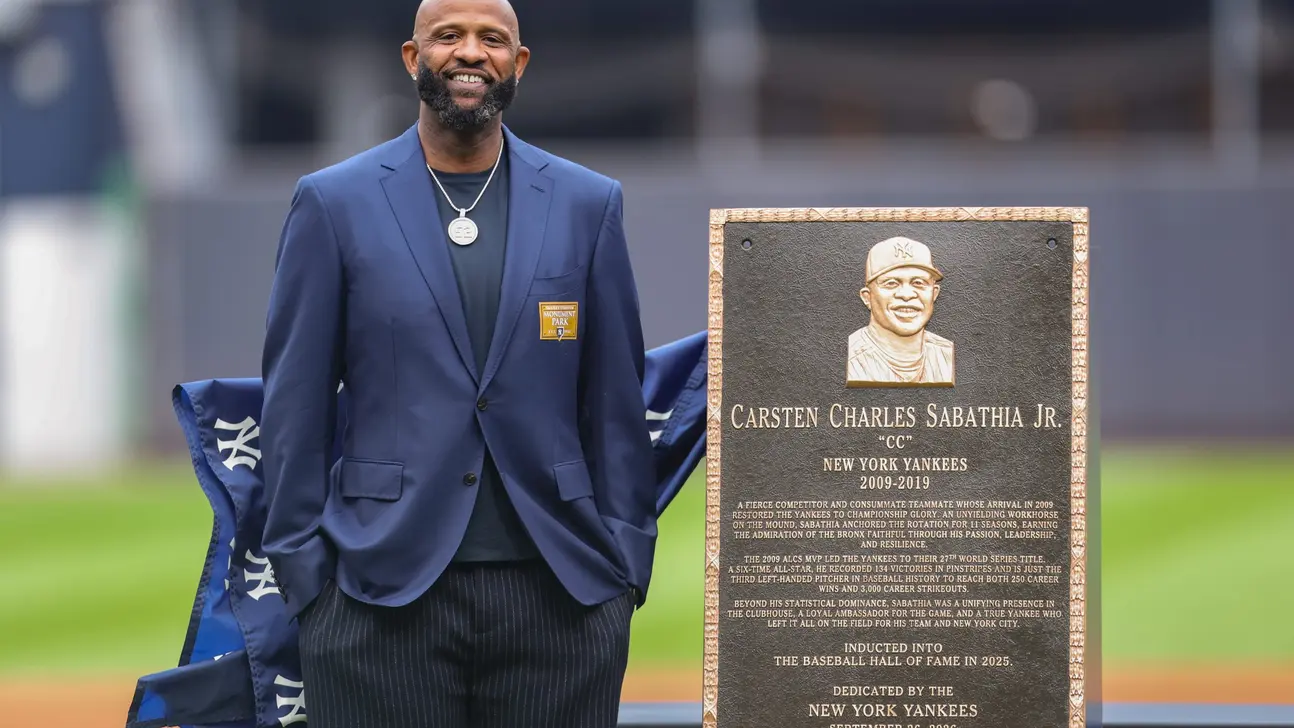 A man in a blue suit stands beside a plaque honoring CC Sabathia and a draped Yankees banner.
