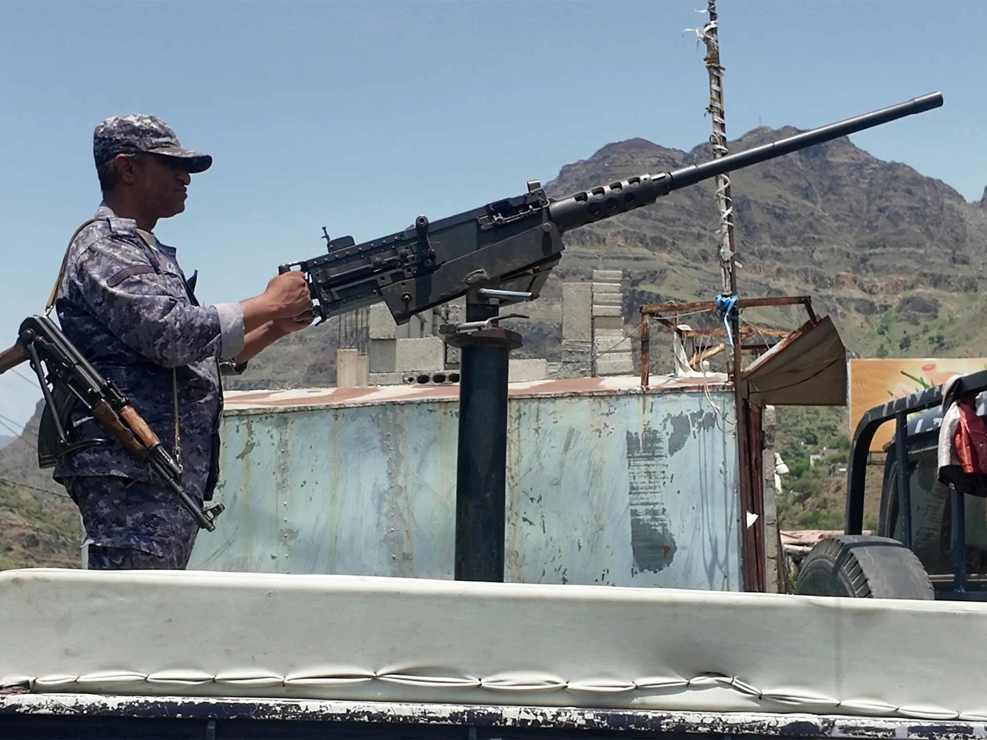 Armed soldier operates a mounted heavy machine gun against a mountainous backdrop.