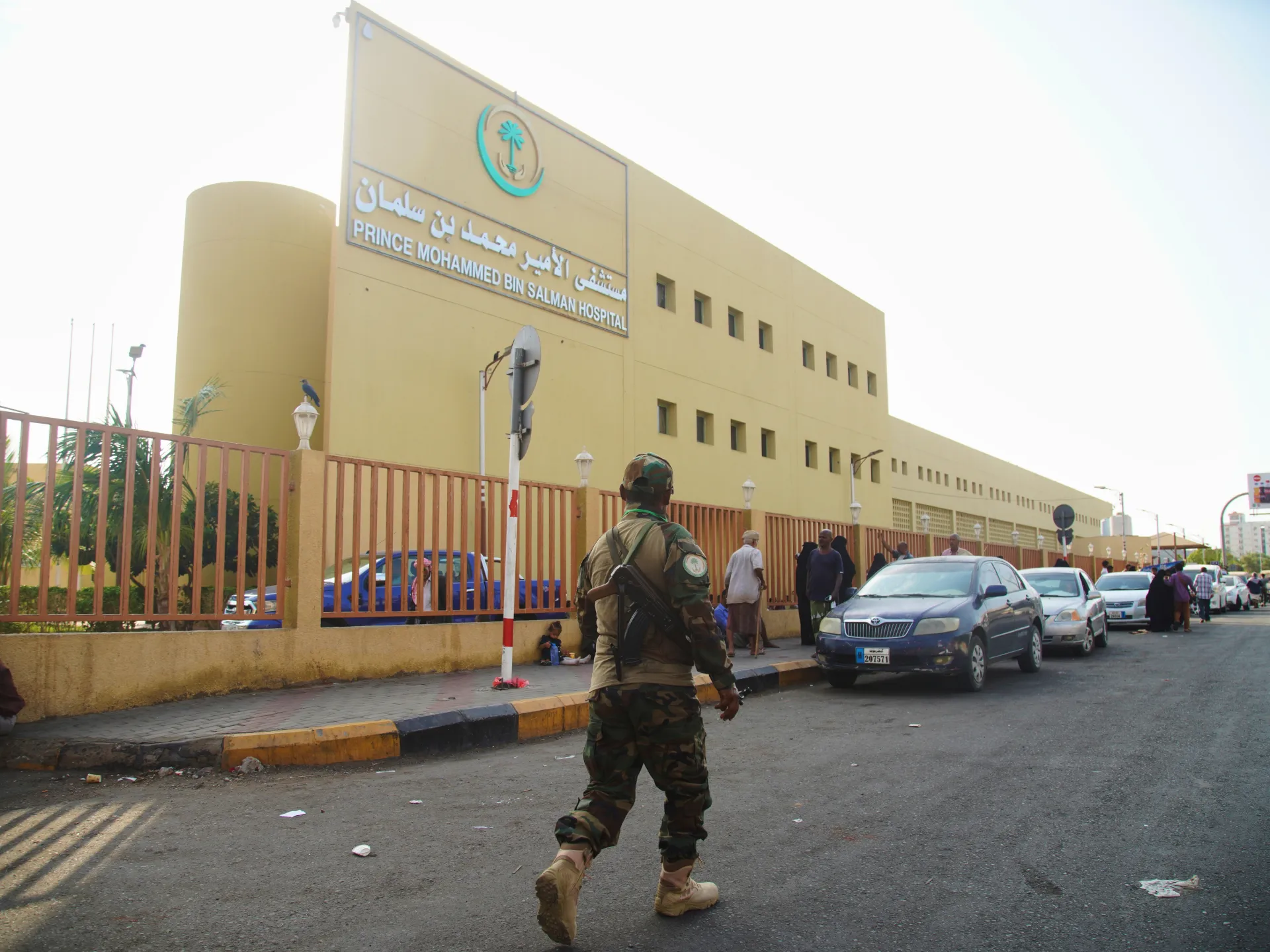 A uniformed man walks toward parked cars outside a large hospital building with Arabic and English signage.