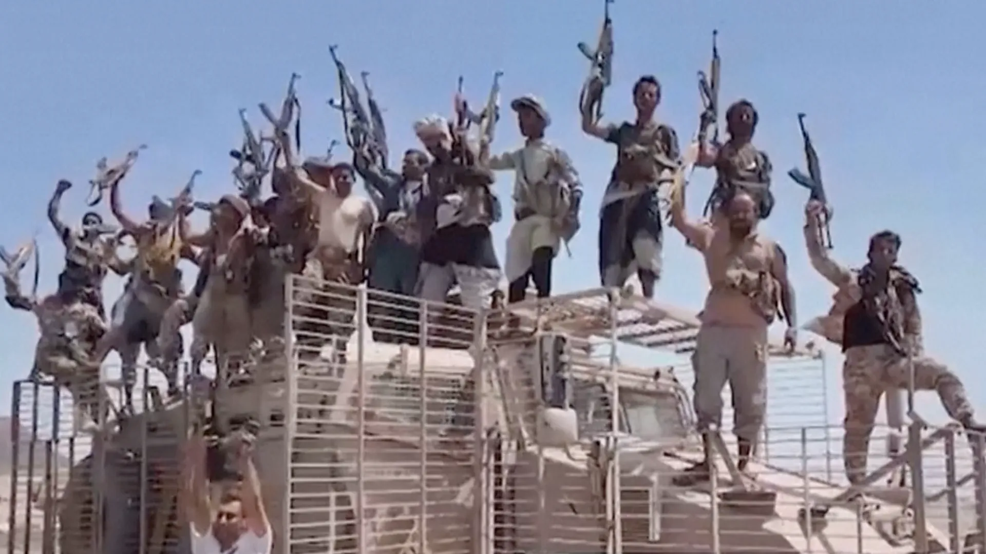 Armed men raise rifles while standing on armored vehicles under a clear blue sky.