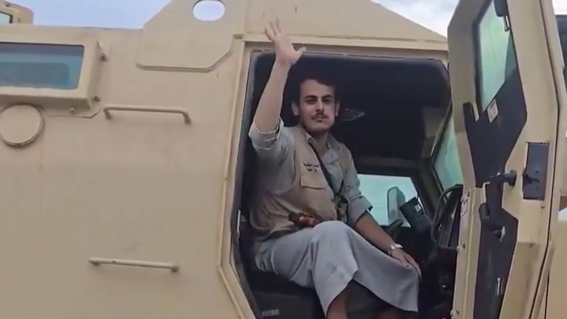 Man in military-style clothing waves while seated inside an armored vehicle.