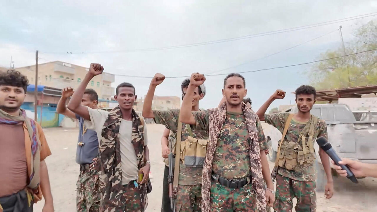 Several men in camouflage raise their fists on a street as a microphone is held toward them.