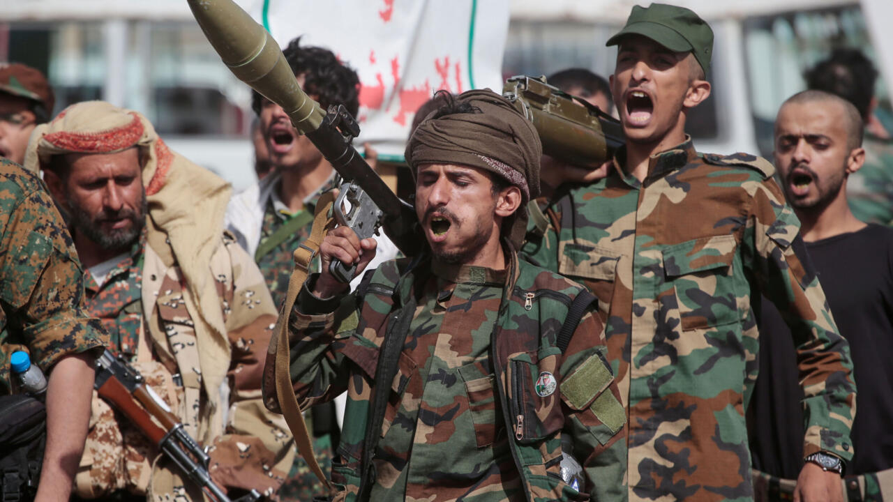 Armed men in camouflage gather outdoors, some shouting and carrying rifles and a shoulder-fired weapon