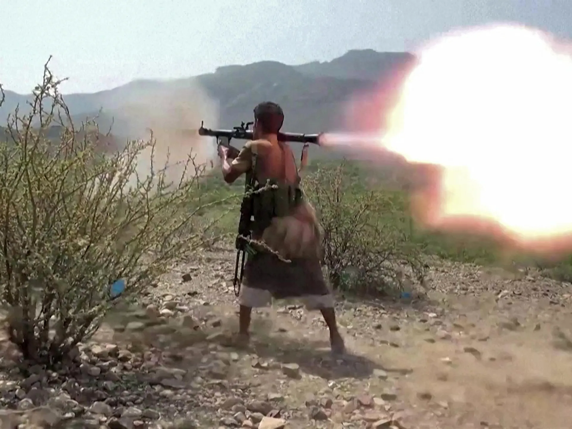 Armed man firing a shoulder-launched weapon amid rocky scrubland and distant mountains