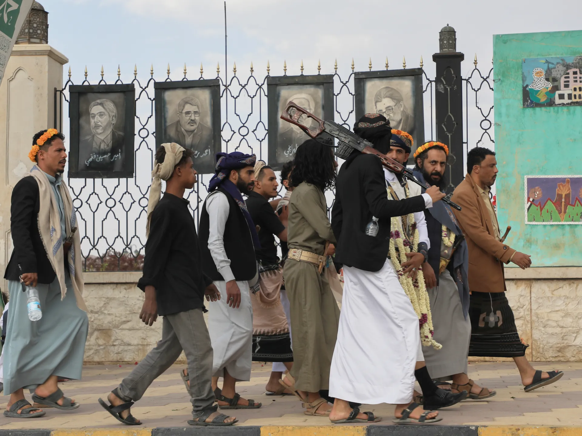 Men walk past portraits and murals, with one carrying a rifle and others wearing traditional clothing.