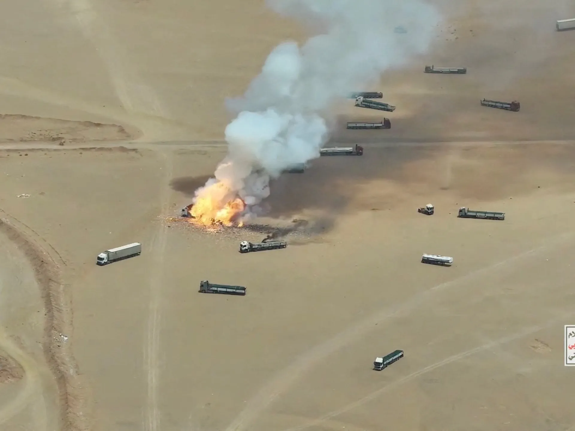 Aerial view of an explosion and thick smoke among trucks on a desert roadway.