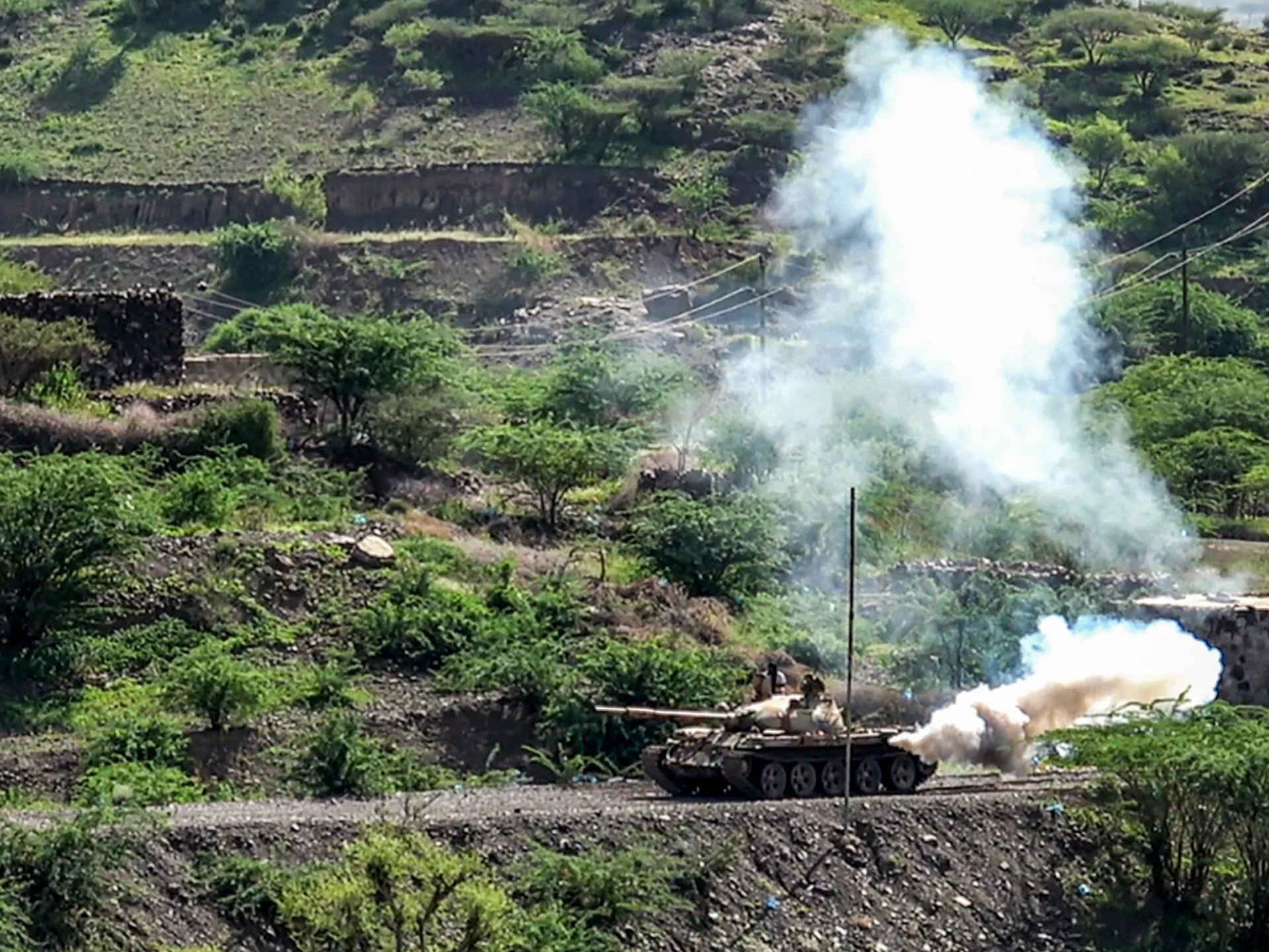 Tank fires across a rugged, green hillside, producing large clouds of white and brown smoke.