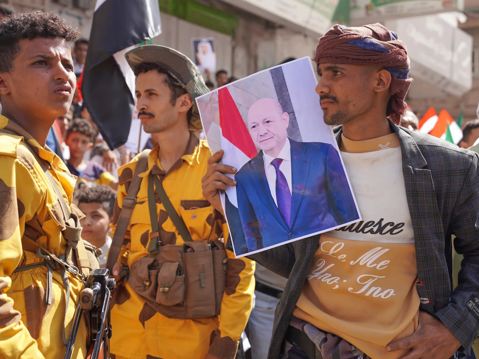 Men in yellow uniforms stand among a crowd as another man holds a portrait poster.