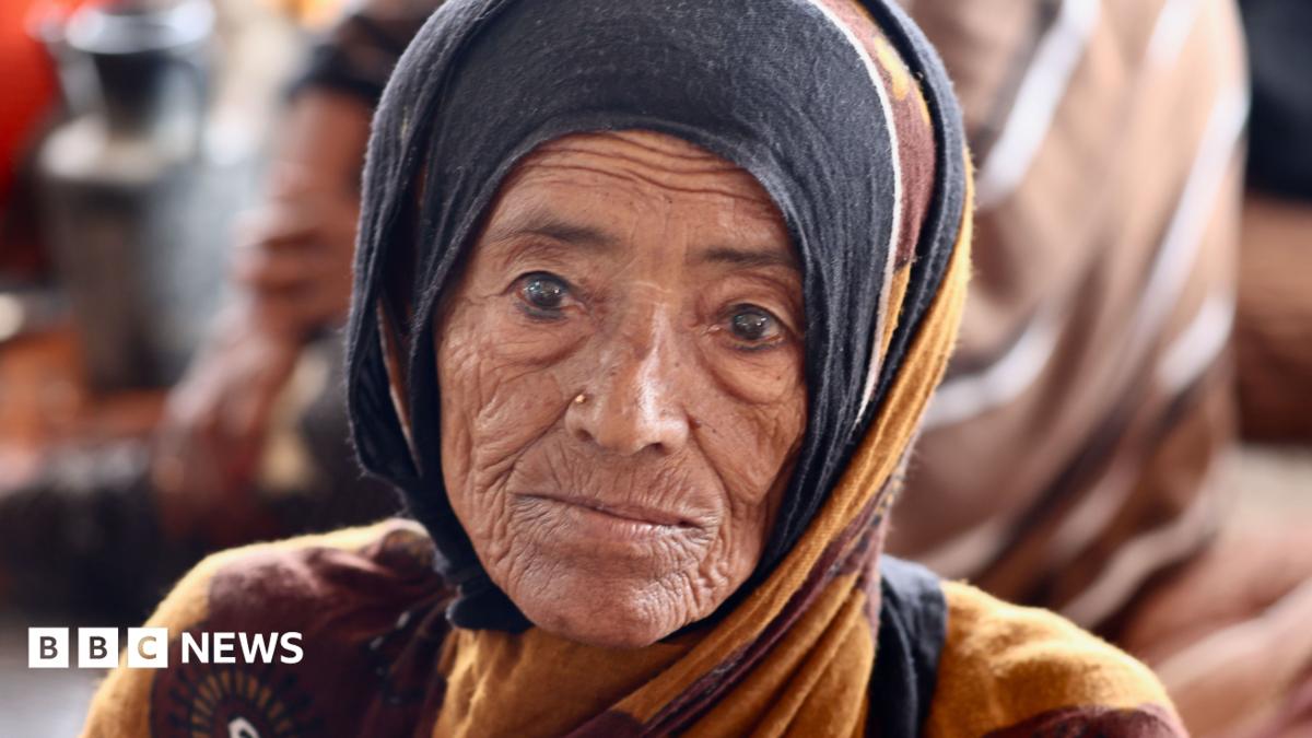 Elderly woman in a black and ochre headscarf looks directly toward the camera.