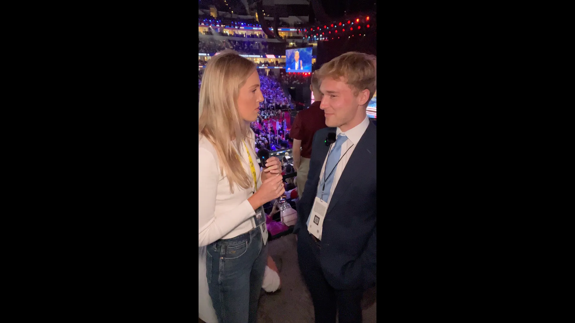 Young woman interviews a suited young man inside a crowded arena during a political event