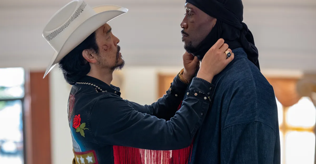 Two men face each other as one adjusts the other's black head covering indoors.