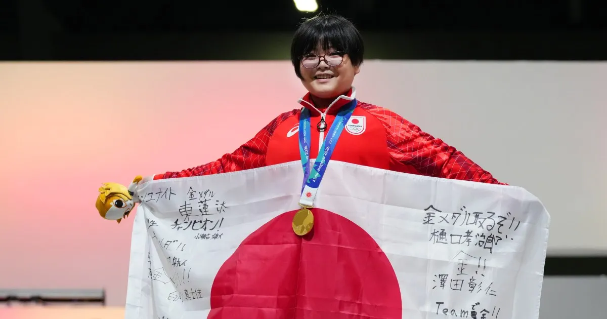 Young athlete in a red jacket holds a Japanese flag and wears a gold medal and glasses.