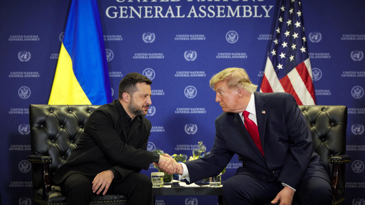 Two men in suits shake hands while seated before Ukrainian and American flags at a United Nations backdrop.