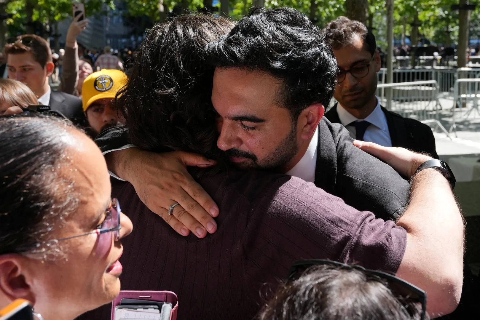 A bearded man in a dark suit embraces another person amid a crowded outdoor gathering.
