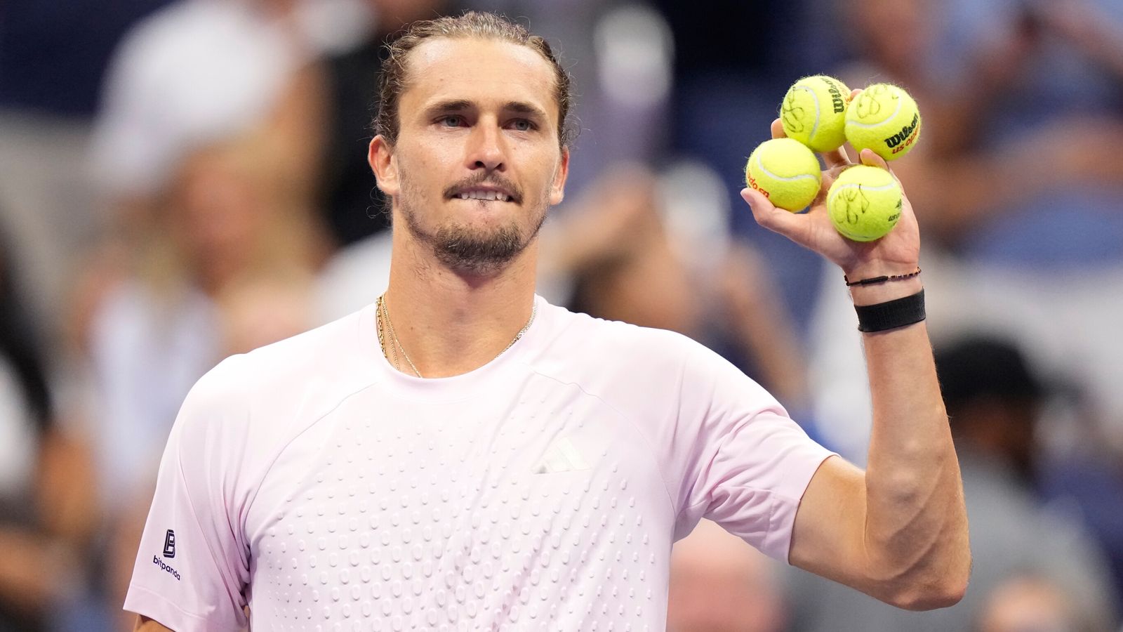 Tennis player holds four balls aloft during a match, with a blurred crowd behind him.