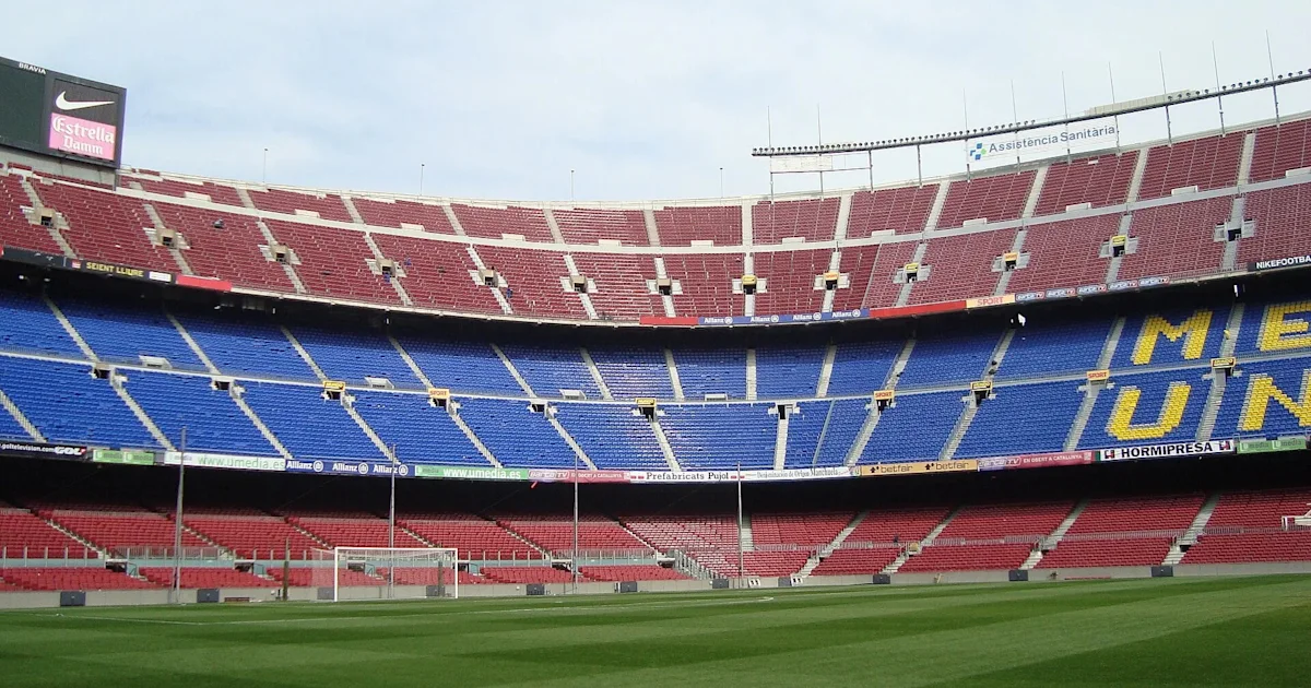Empty football stadium with blue and red seating, a green pitch, and yellow lettering across the stands.