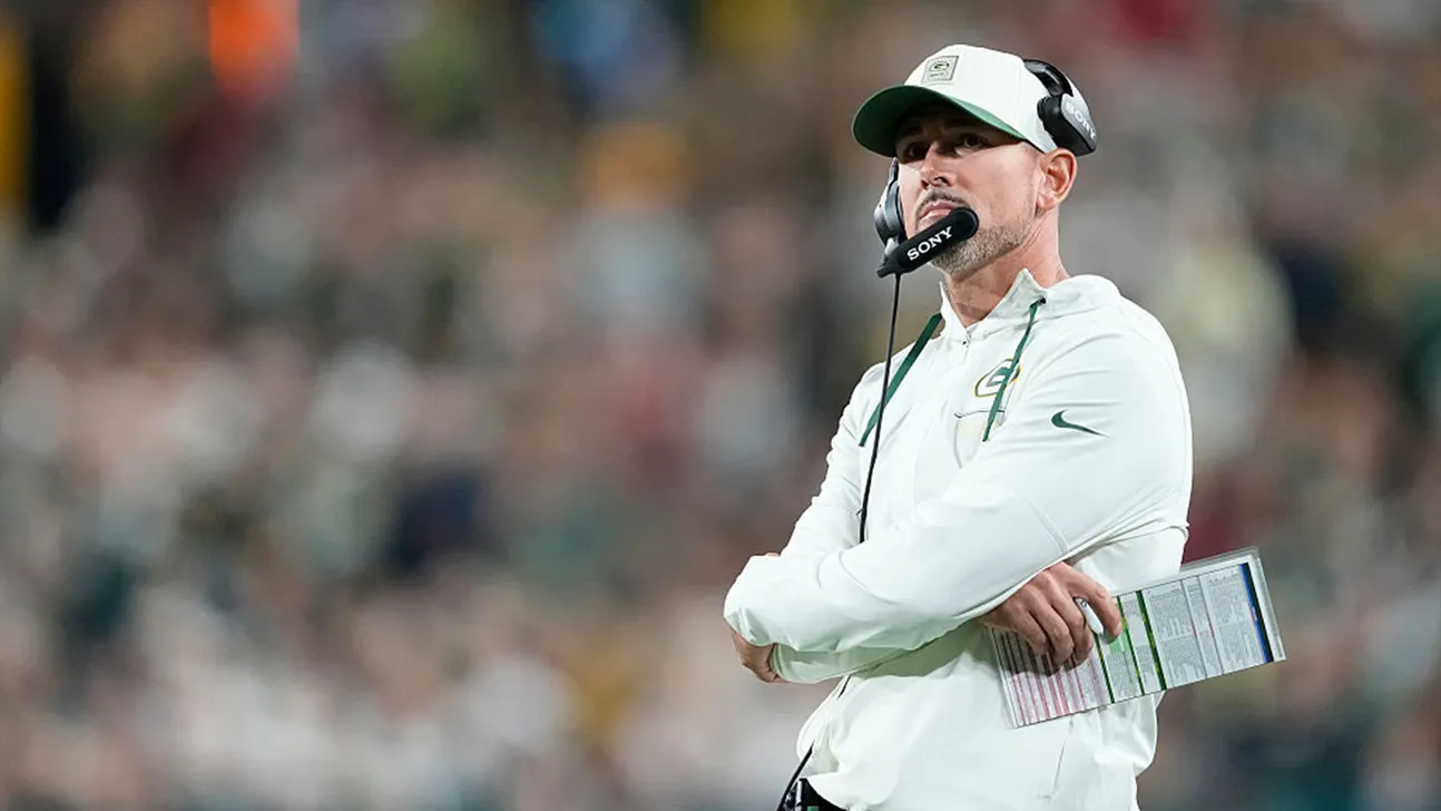 A football coach in a white cap and pullover stands with arms crossed, holding a play sheet.