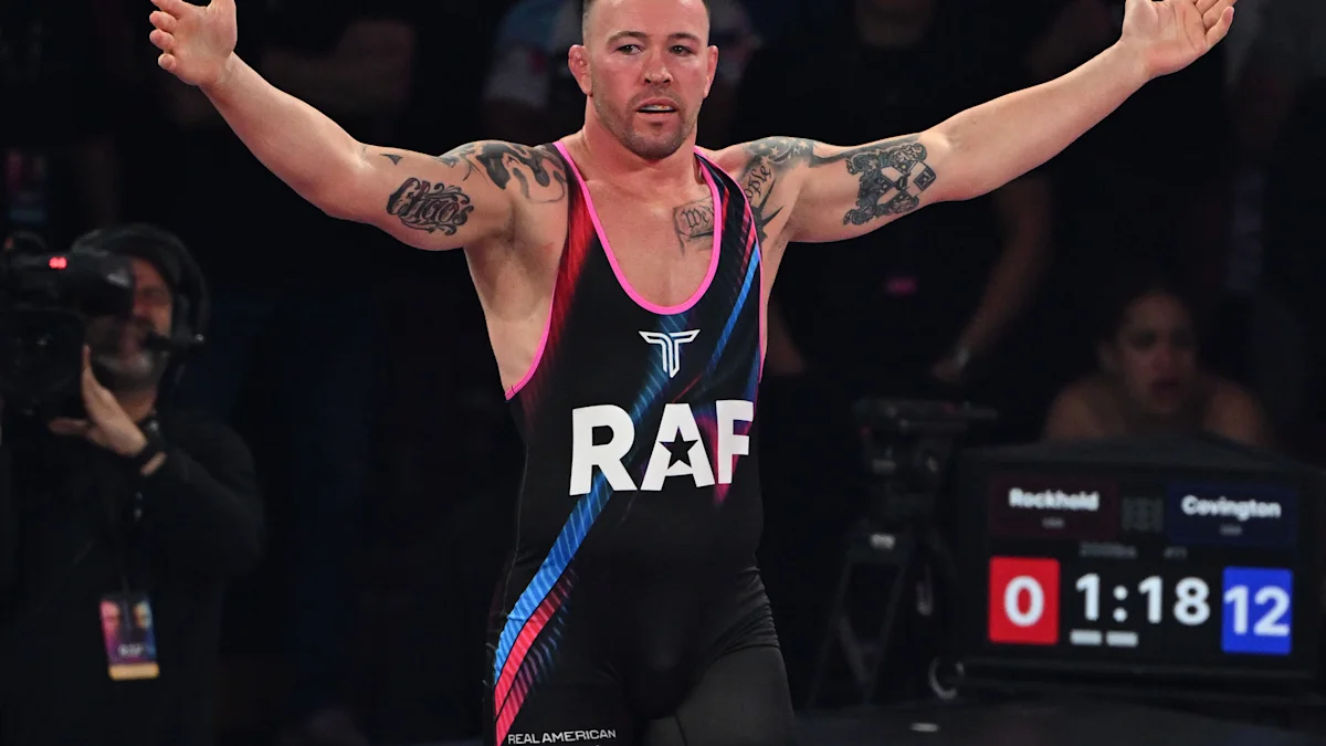 Wrestler in a black singlet raises both arms beside a match scoreboard.
