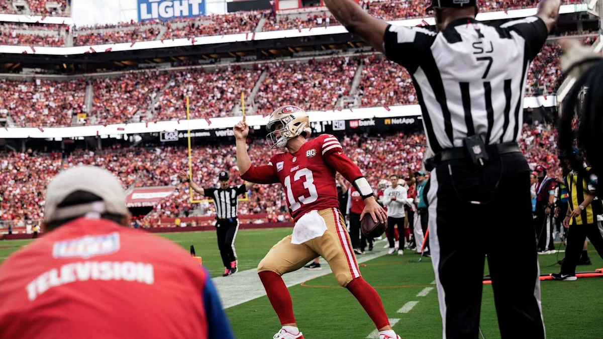 A football player in a red jersey raises one finger as a referee signals beside the field.