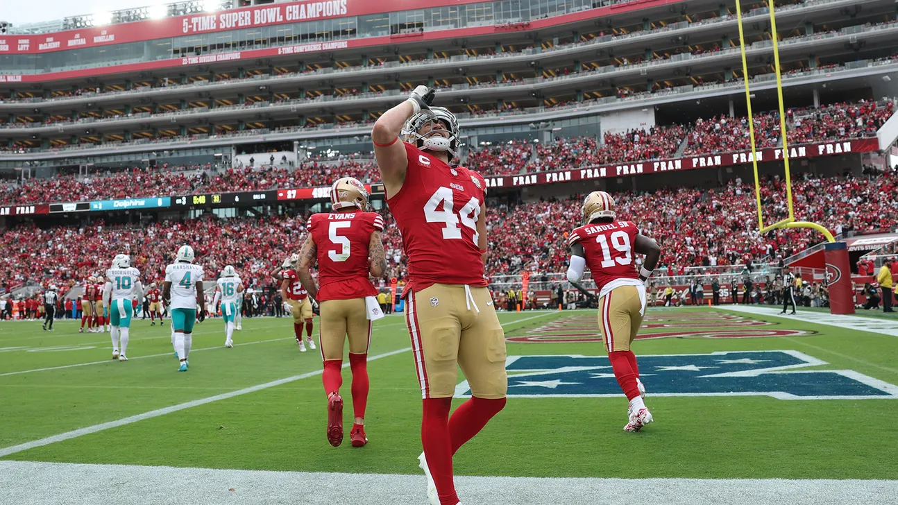 A 49ers player in a red number 44 jersey stands on the field as teammates walk away.