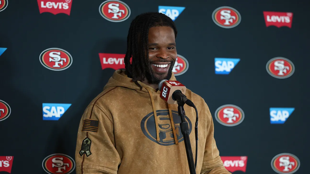 A smiling man in a 49ers hoodie speaks into a microphone before a team-branded backdrop.