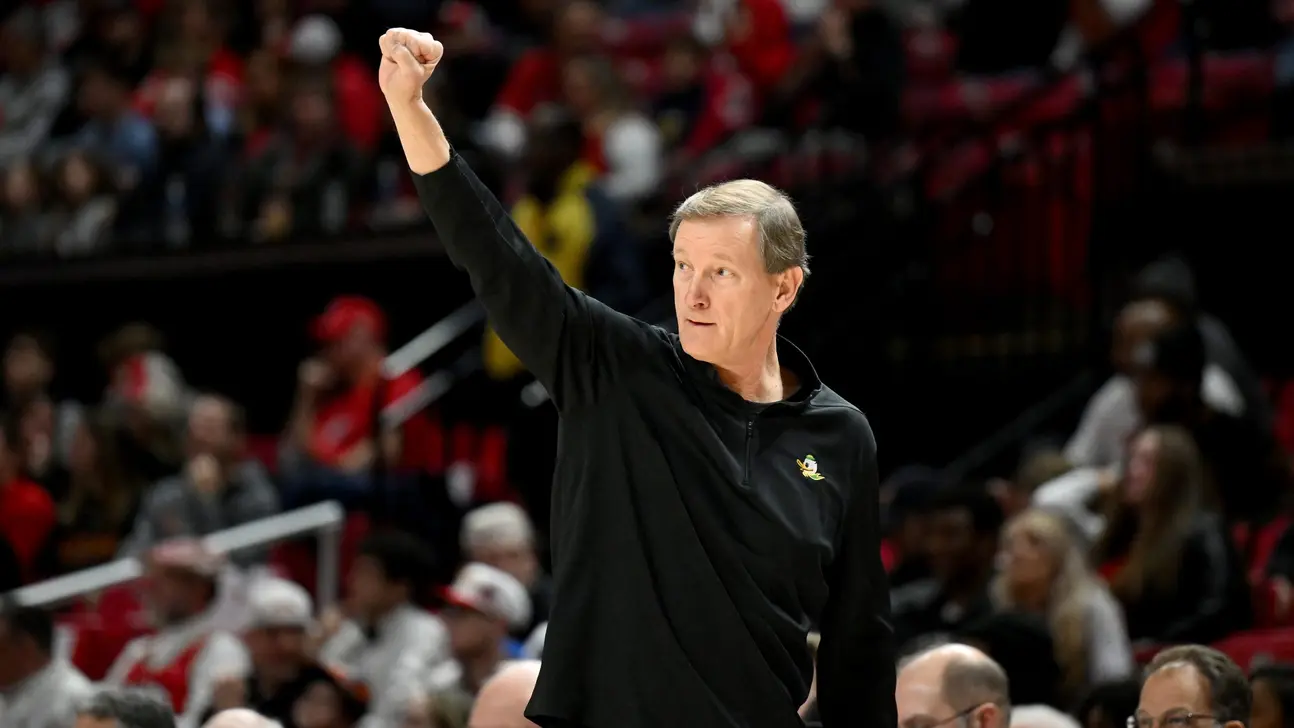 Basketball coach in a black pullover raises his fist during a game as spectators watch.