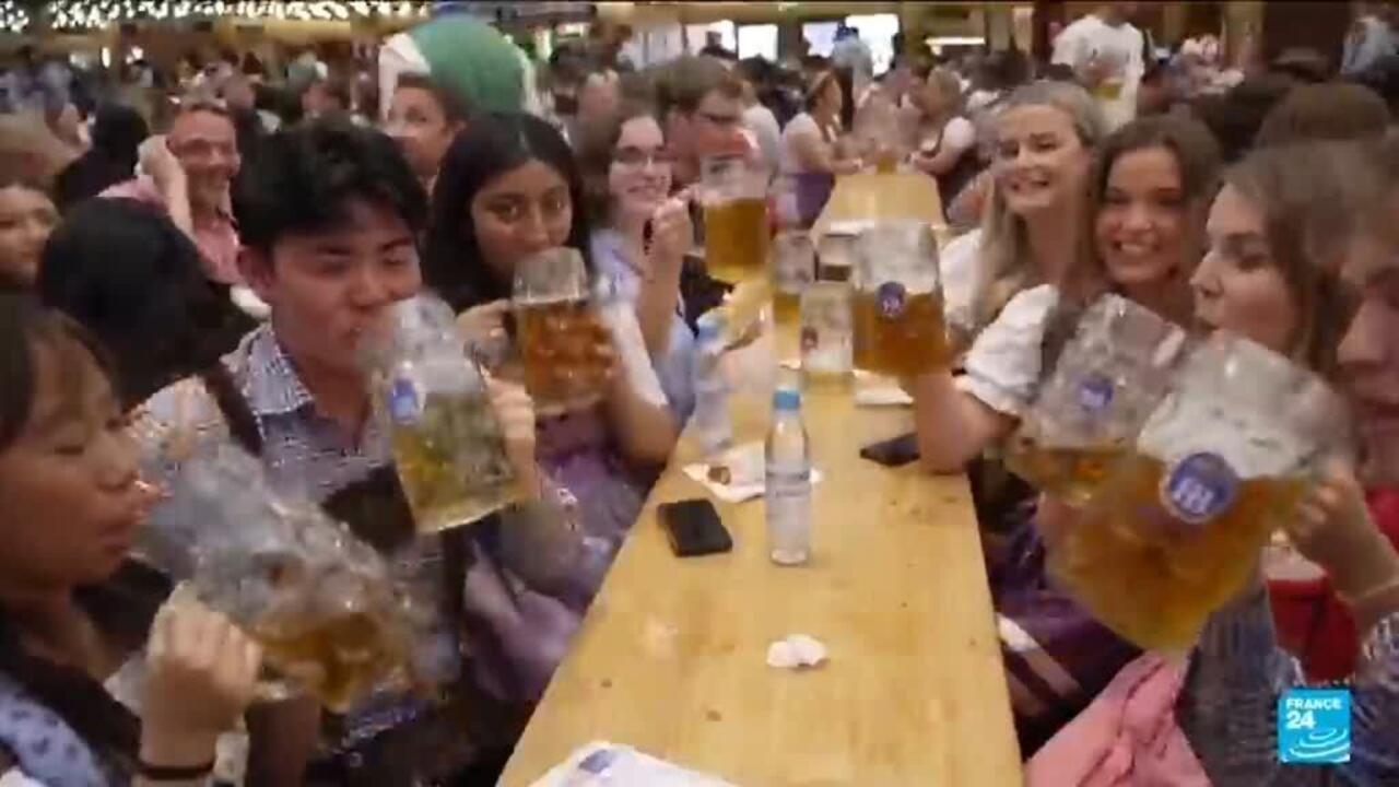 Festivalgoers raise oversized glass beer mugs around a crowded wooden table in a busy hall.