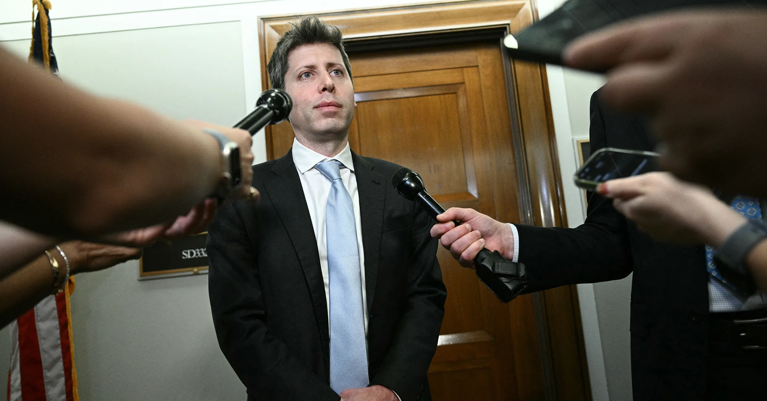 Man in a dark suit stands as reporters extend microphones and a phone toward him.