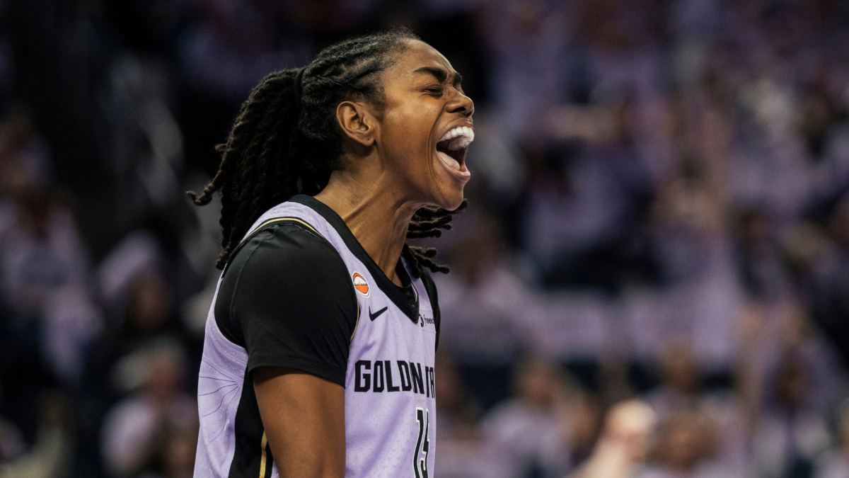 Basketball player in a light purple jersey shouts during a game, with a blurred crowd behind her.