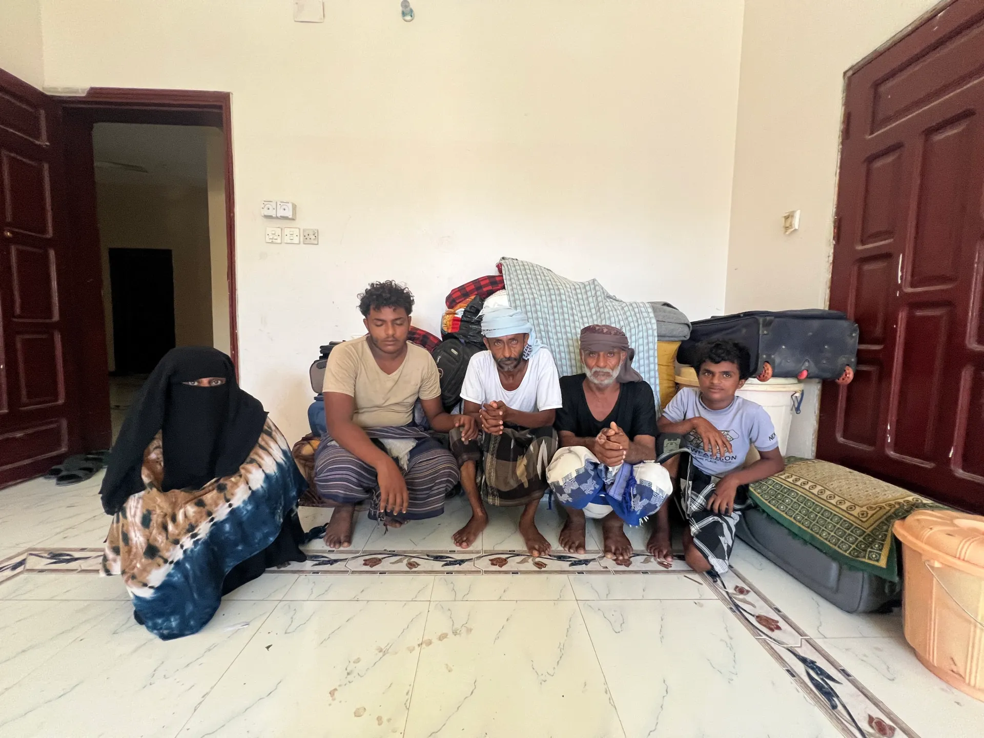 Five people sit on a tiled floor beside stacked bags, bedding, and suitcases in a room.